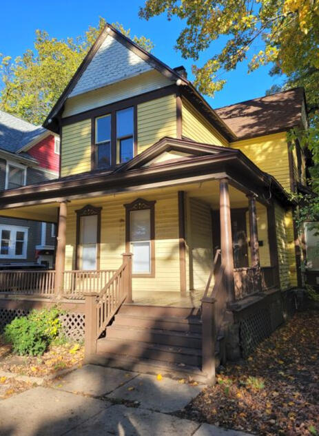 A yellow house with brown trim and a front porch. There are leaves on the ground and a cloudless blue sky.